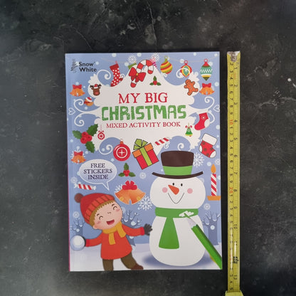 A children's activity book titled 'My Big Christmas Mixed Activity Book' by Snow White, featuring a colorful winter illustration with a snowman, child, ornaments, and a yellow measuring tape standing beside it on a dark textured surface.