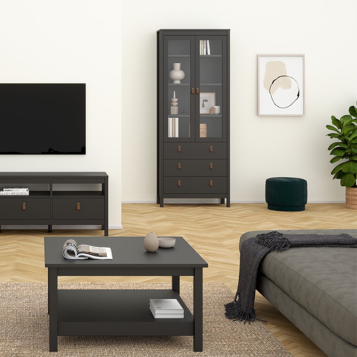 A staged living room scene showing a tall Barcelona black glass-front cabinet with drawers, a matching dark coffee table, a gray sofa, a TV stand, a plant, and a green ottoman against a light wall.
