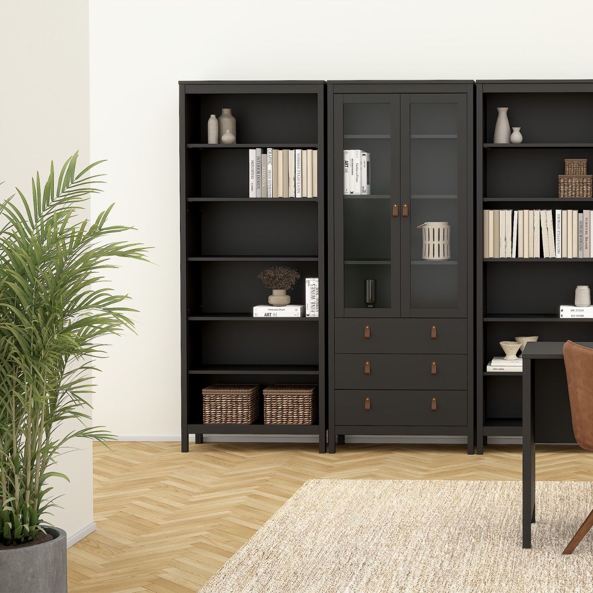 A three-piece black wooden bookcase and cabinet set styled in a modern living room with light wood herringbone floors, a beige rug, and a potted plant.