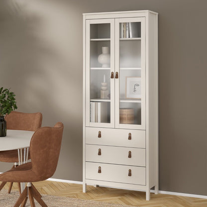 A tall Barcelona glass-front cabinet with three lower drawers and small brown metal pulls, shown in a styled room with a dining table and chairs against a taupe wall.