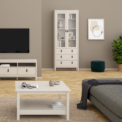 A staged living room scene showing a tall cashmere glass-front cabinet with drawers, a matching low media console, a coffee table, a sofa, wall art, a plant, and a dark green ottoman on a wood floor.