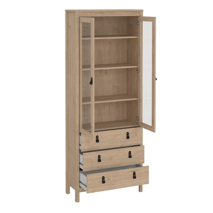 A tall light oak wood storage cabinet with a glass-front upper door, open to reveal shelves, and three drawers below with black metal handles.