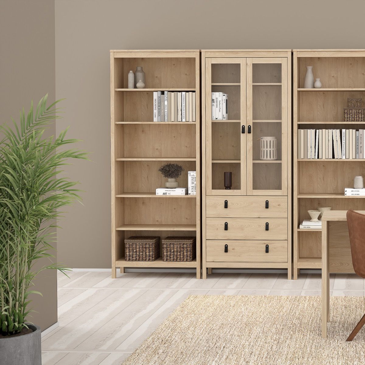 A light natural wood bookcase and storage unit with open shelves, glass-front upper doors, and drawers, shown in a styled room with a potted plant, rug, and part of a chair.