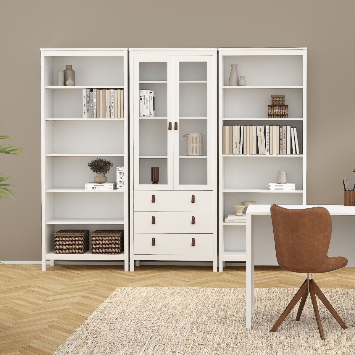 A set of three white modular Barcelona bookcases and drawers, styled with books, vases, and baskets, positioned in a modern living room setting next to a brown chair and a white desk.