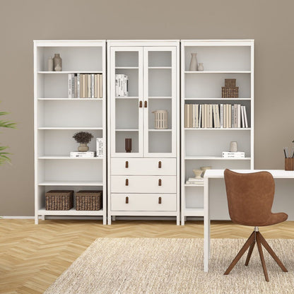 A set of three white modular Barcelona bookcases and drawers, styled with books, vases, and baskets, positioned in a modern living room setting next to a brown chair and a white desk.