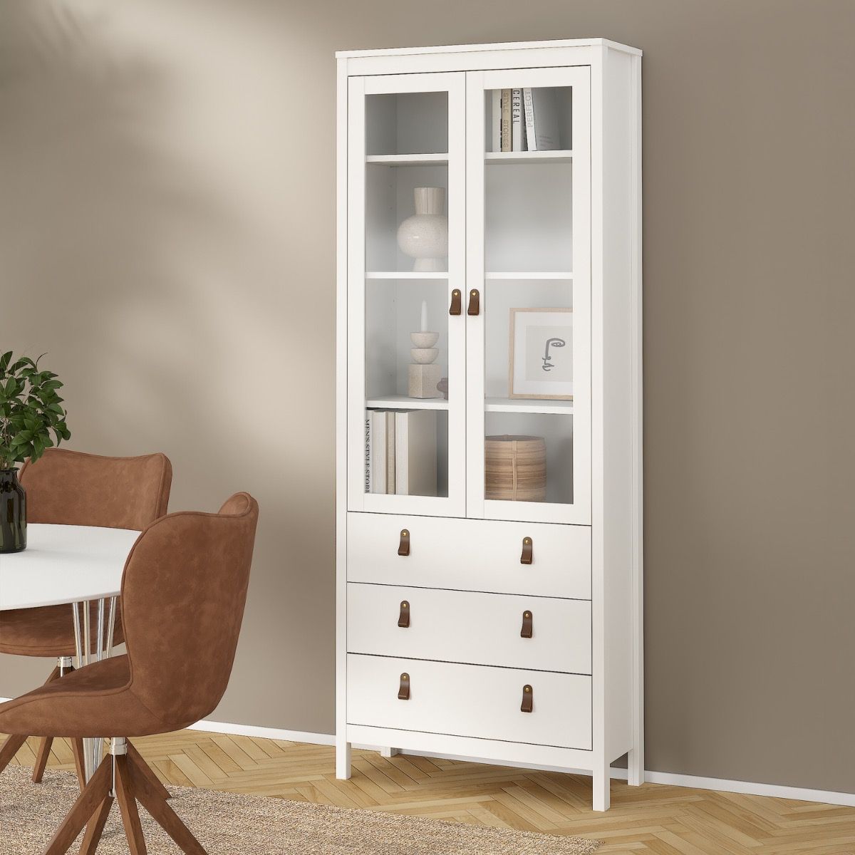 A tall white wooden Barcelona display cabinet with two glass-front upper doors and three lower drawers, styled in a dining room next to a table and chairs against a beige wall.