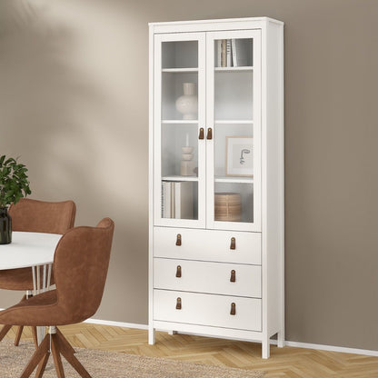 A tall white wooden Barcelona display cabinet with two glass-front upper doors and three lower drawers, styled in a dining room next to a table and chairs against a beige wall.