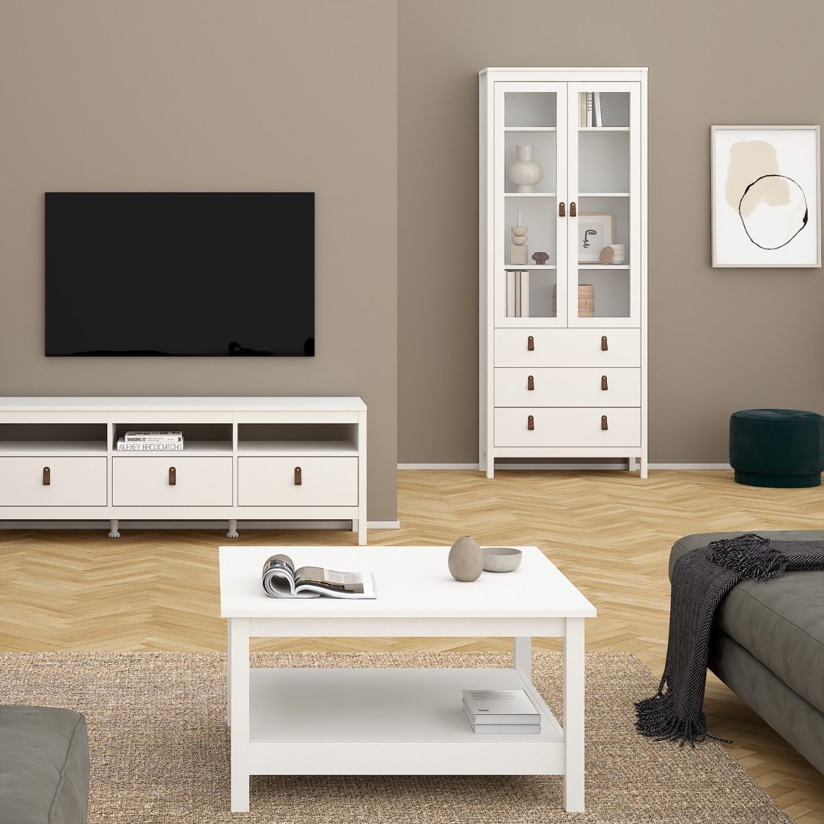 A modern living room setup featuring a white coffee table in the foreground, a white TV stand and matching wall cabinet in the background, set against warm taupe walls and light wood herringbone flooring.