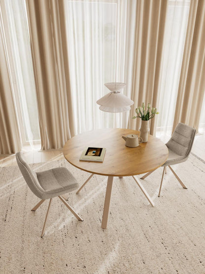 A round light wood dining table with three light gray upholstered swivel chairs in a bright, modern room with beige curtains and sheer white window drapes.