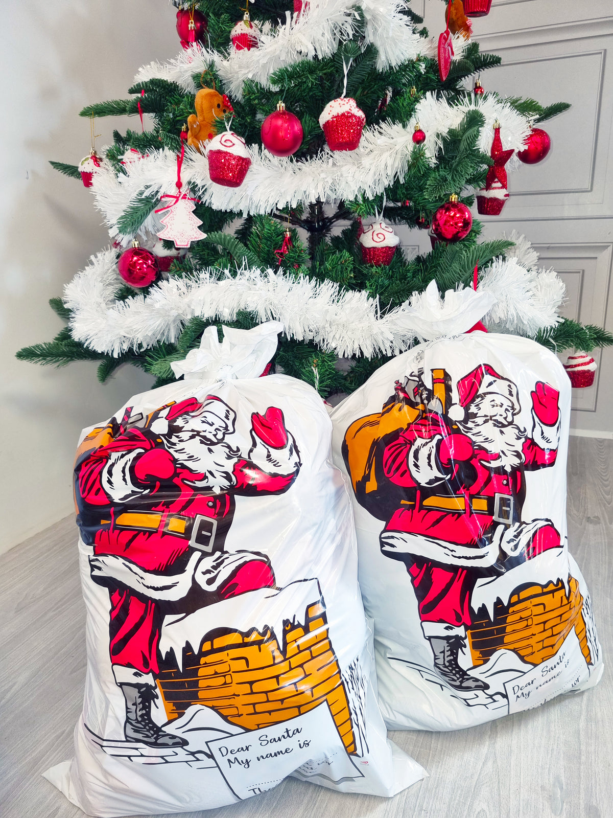 Two large white Christmas gift bags printed with a vintage-style illustration of Santa Claus carrying a sack, placed in front of a decorated Christmas tree in an indoor room.