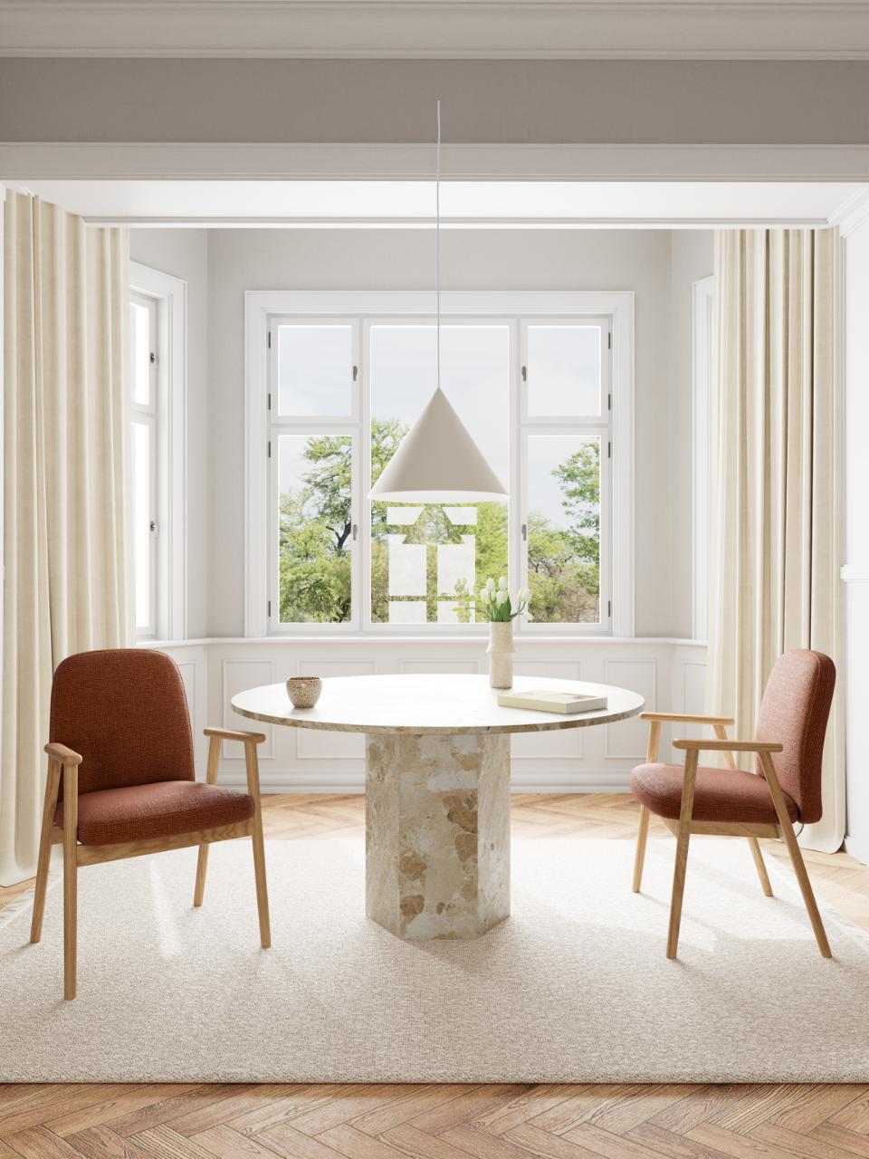 A modern dining setup in a bright, sunlit room featuring a round white marble-top table with a textured stone pedestal base, flanked by two mid-century style armchairs with brown upholstery and light wood frames, under a conical pendant light.
