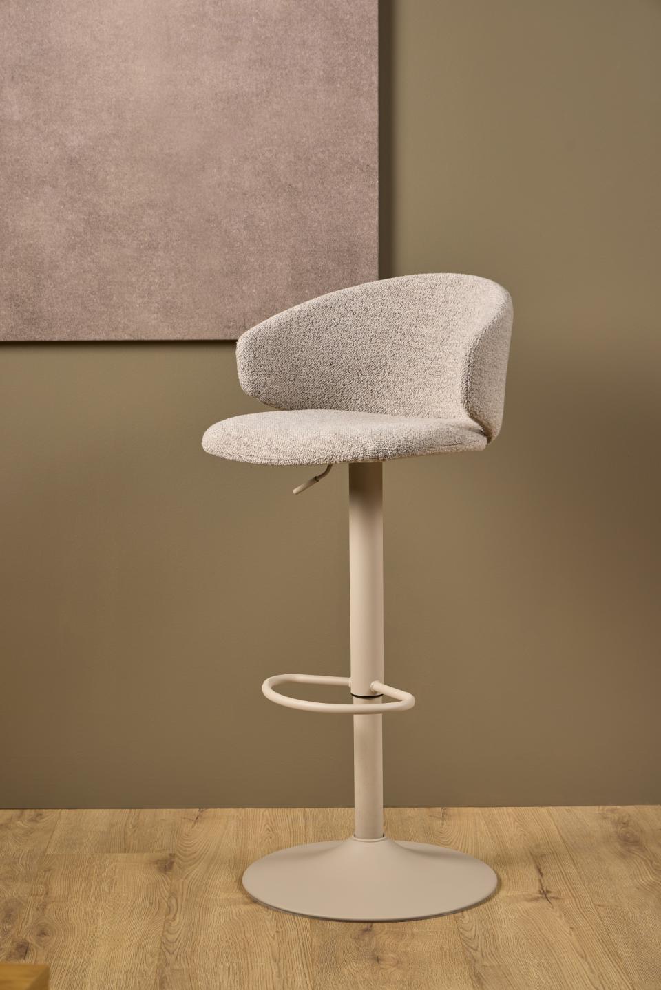 A modern swivel bar stool with a textured light gray upholstered seat and curved backrest, mounted on a slim cream-colored metal pedestal base with a footrest ring, set against a warm brown wall and light wood floor.