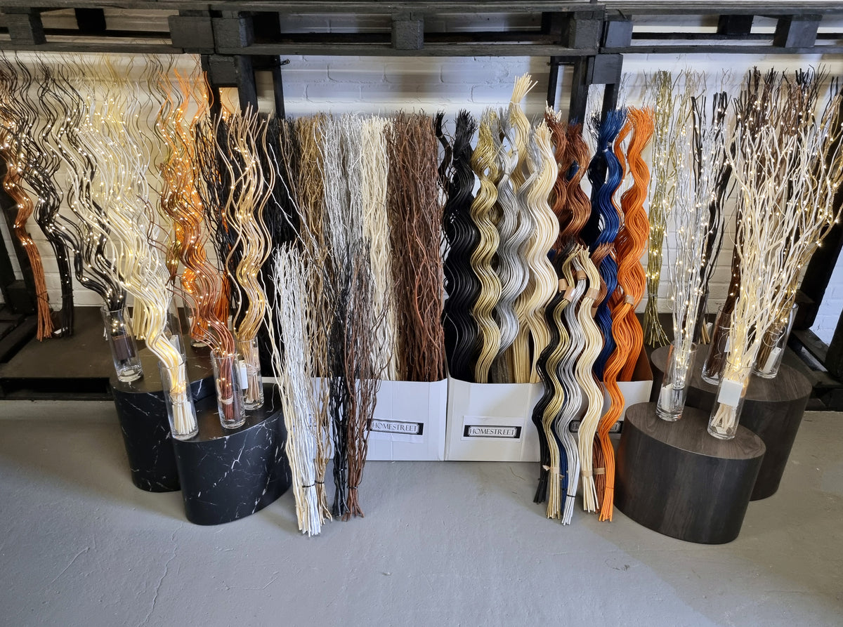 A display of decorative wavy branches in multiple colors, including cream, gold, brown, black, orange, and blue, arranged on black and white pedestals in a Homestreet showroom.