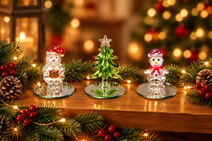 Three small Christmas-themed glass ornaments displayed on a wooden surface: two snowman figures with red hats and scarves flanking a green tree with a star topper, surrounded by pine branches, berries, and warm holiday lights.