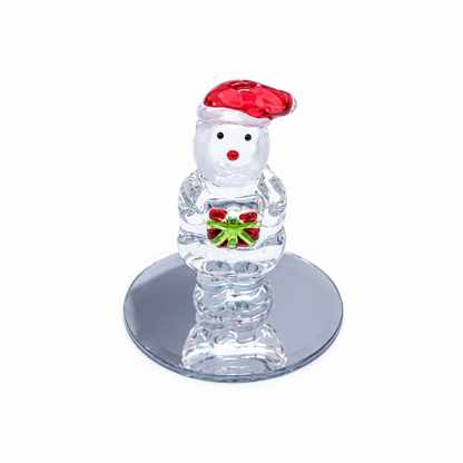 A clear glass Santa Claus figurine wearing a red Santa hat and holding a green gift box with red and white accents, standing on a round mirrored base against a white background.