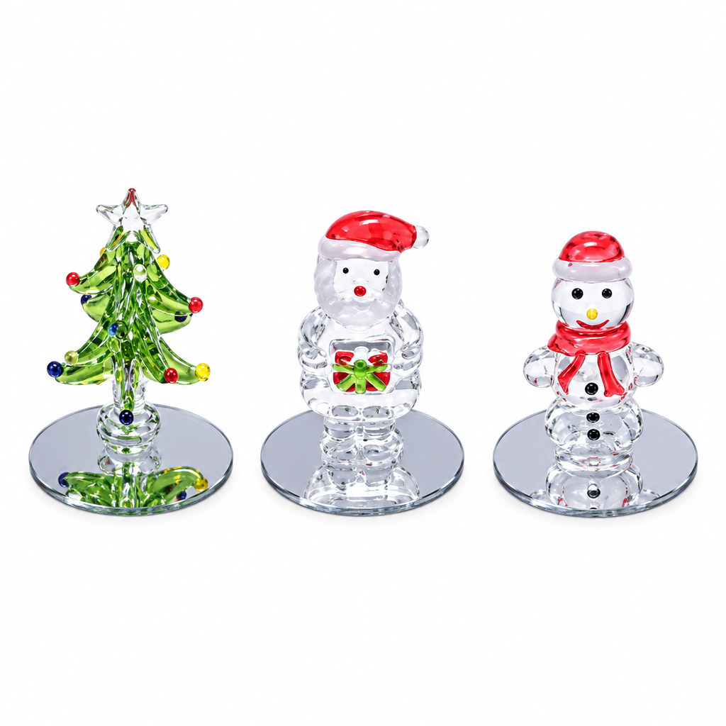 Three clear glass Christmas ornaments on mirrored bases: a green tree with colored accents, a snowman in a red Santa hat holding a gift, and a snowman in a red Santa hat and scarf.