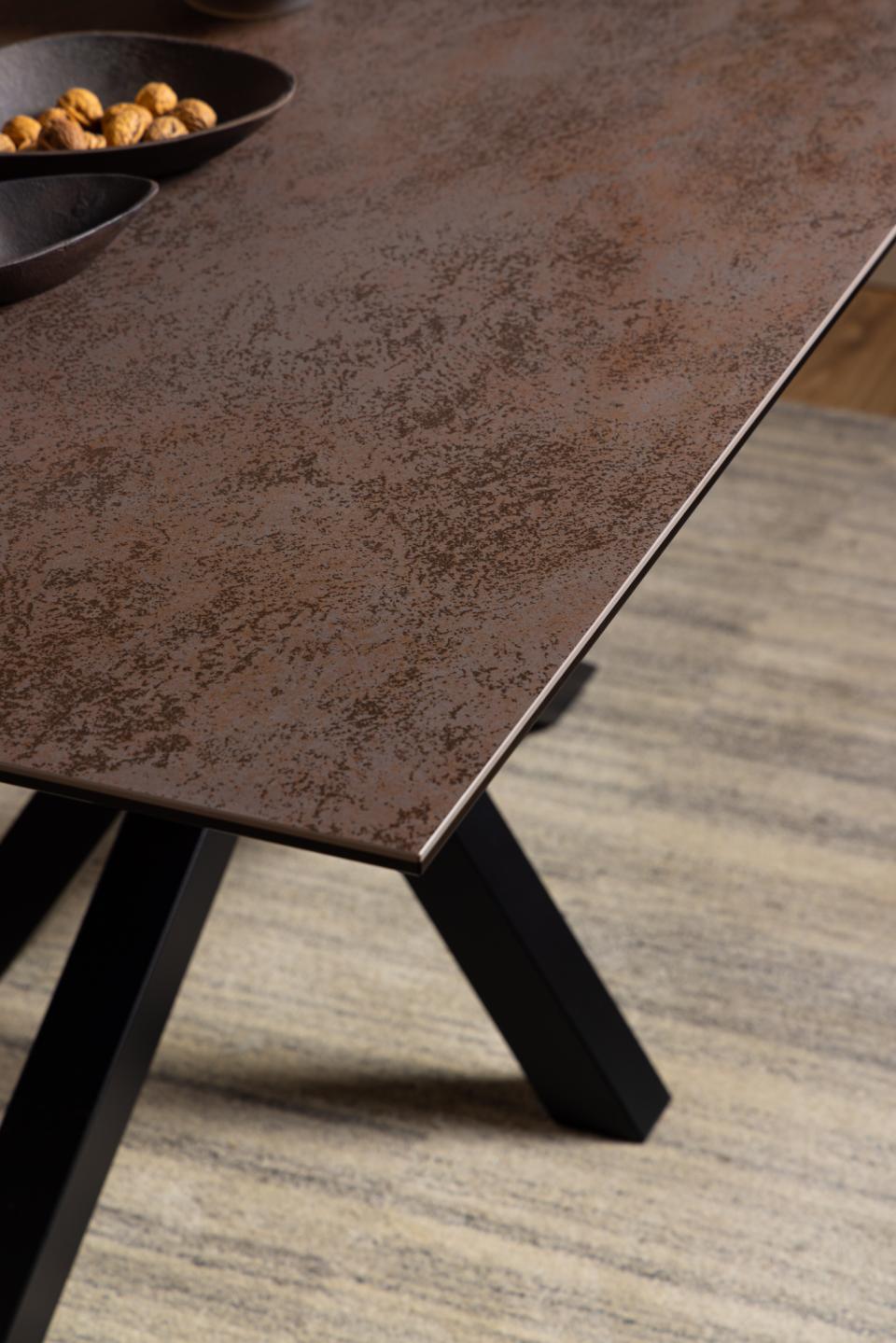 Close-up of a rectangular table with a dark brown ceramic top and black angled legs, shown on a gray wood floor with a bowl of nuts in the corner