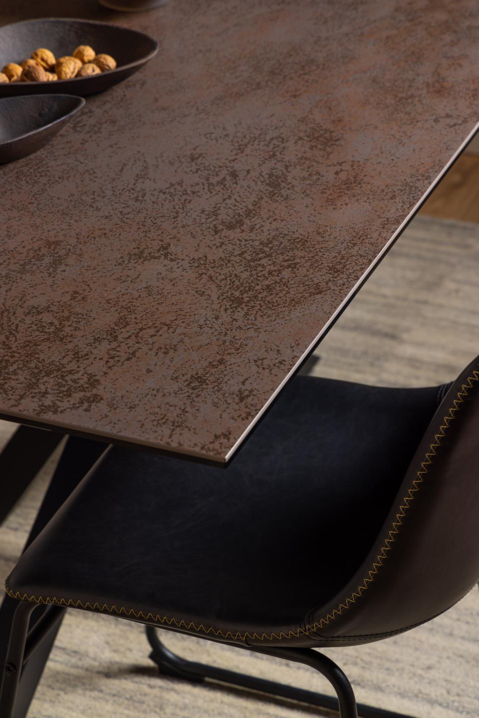 Close-up of a dark brown textured dining table corner with a black upholstered chair underneath, set in a modern interior with wood flooring and a bowl of nuts in the upper left.