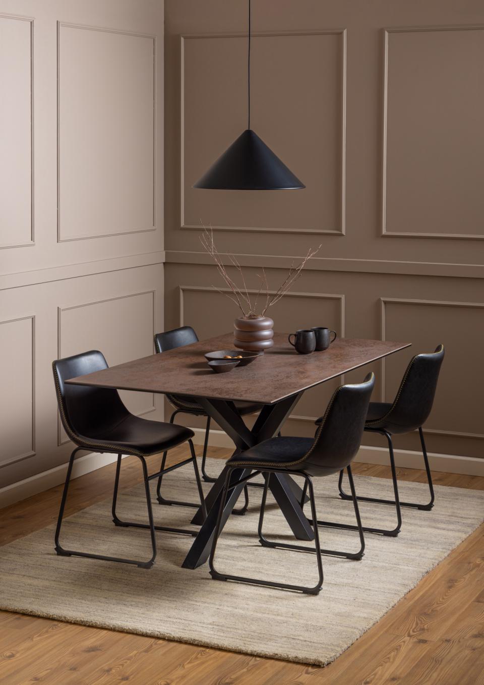 A dining set featuring a rectangular ceramic table with a black metal X-base and six black chairs, styled in a modern dining room with beige paneled walls and a light rug.
