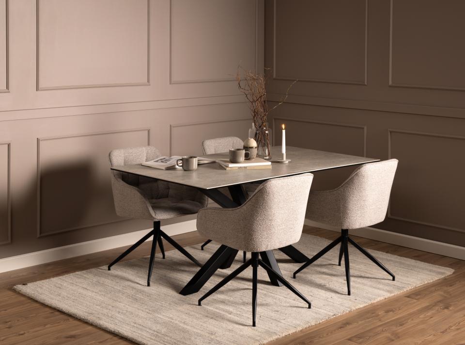 A modern dining set featuring a rectangular light gray ceramic table with a black metal base and four upholstered swivel chairs in a warm beige fabric, set on a textured rug in a dining room with brown paneled walls.