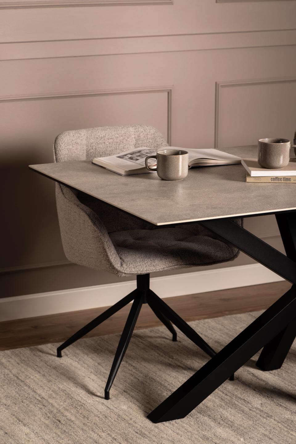 A modern dining table with a grayceramic top and black metal X-shaped legs, paired with a textured light gray upholstered swivel chair. The scene is styled with a mug, an open book, and a stack of books on the tabletop, set against a beige paneled wall and textured rug.