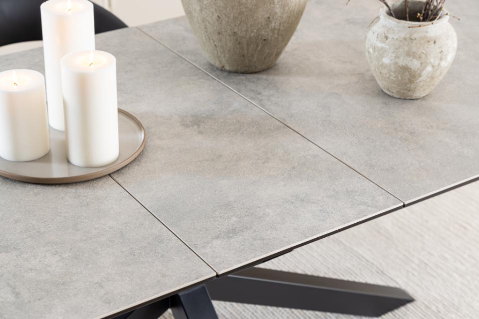 A close-up of a modern rectangular table with a large gray ceramic top and a black metal X-style base, styled with white pillar candles and textured ceramic vases.