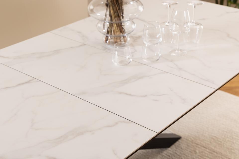 A close-up of a white marble-look rectangular table with thin gray veining, shown with clear glassware and a glass vase on top in a bright indoor setting.
