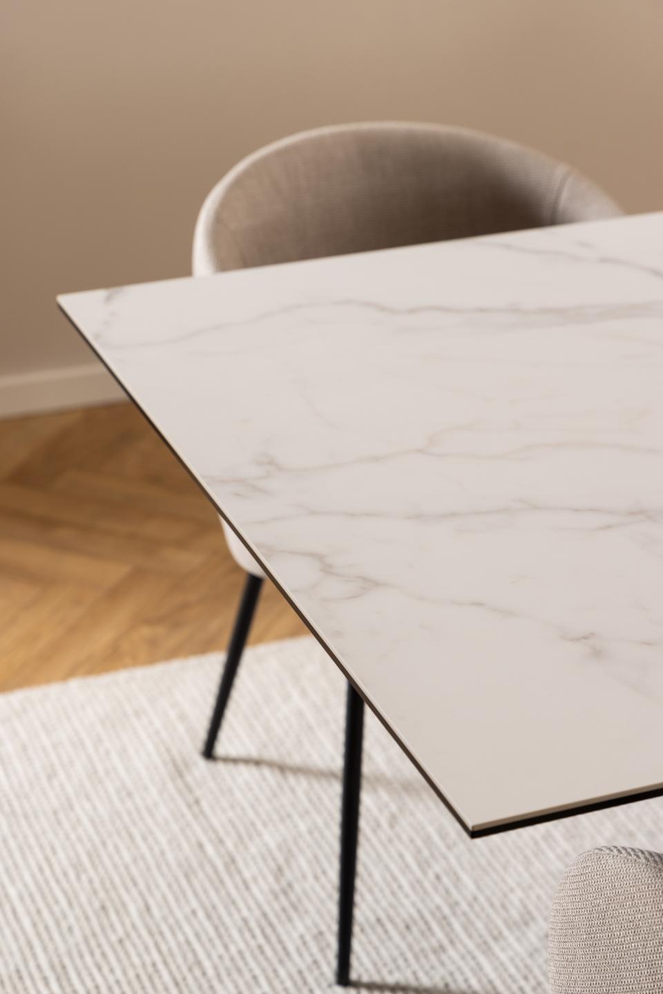 A close-up of a rectangular dining table with a white marble-look top featuring soft gray veining, supported by slim black metal legs, shown in a modern dining room setting with a gray upholstered chair.