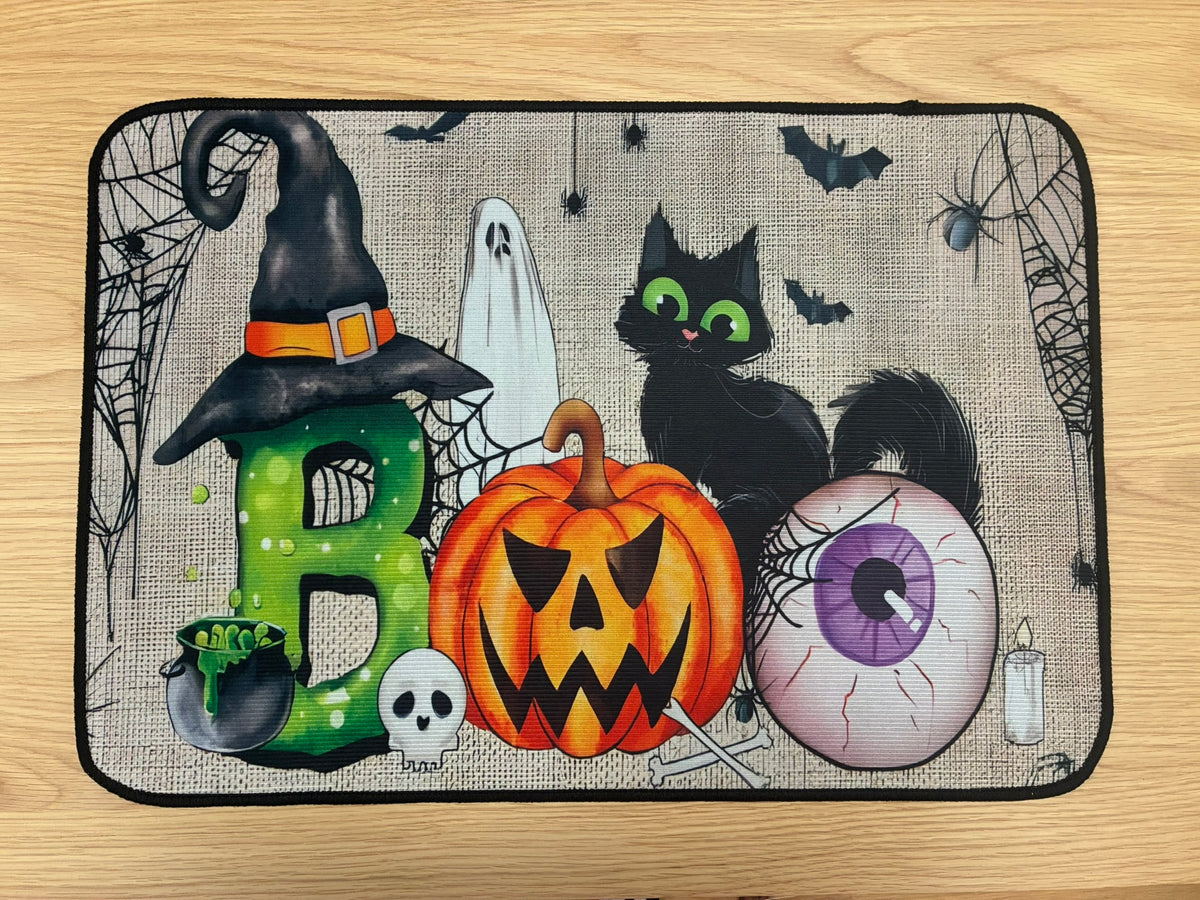 A rectangular Halloween-themed doormat with a black border, featuring a spooky illustration of a green letter B wearing a witch hat, a jack-o'-lantern pumpkin, a black cat with green eyes, a ghost, a eyeball, bats, and cobwebs on a beige burlap-style background, placed on a light wood floor.