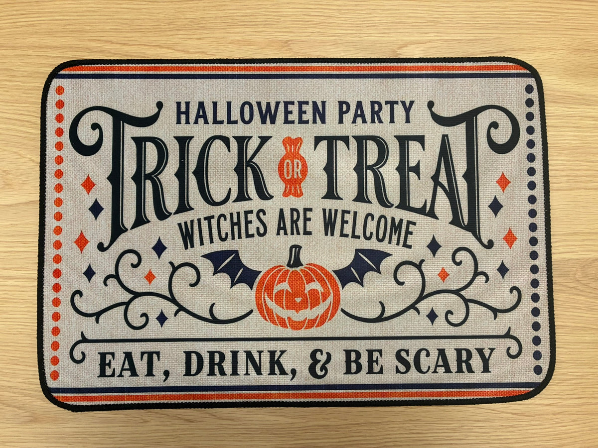 A rectangular Halloween-themed doormat with rounded corners, featuring the text 'HALLOWEEN PARTY TRICK OR TREAT WITCHES ARE WELCOME EAT, DRINK, & BE SCARY' in black lettering, an orange jack-o'-lantern, bats, and decorative scrollwork on a light beige background, photographed on a light wood floor.