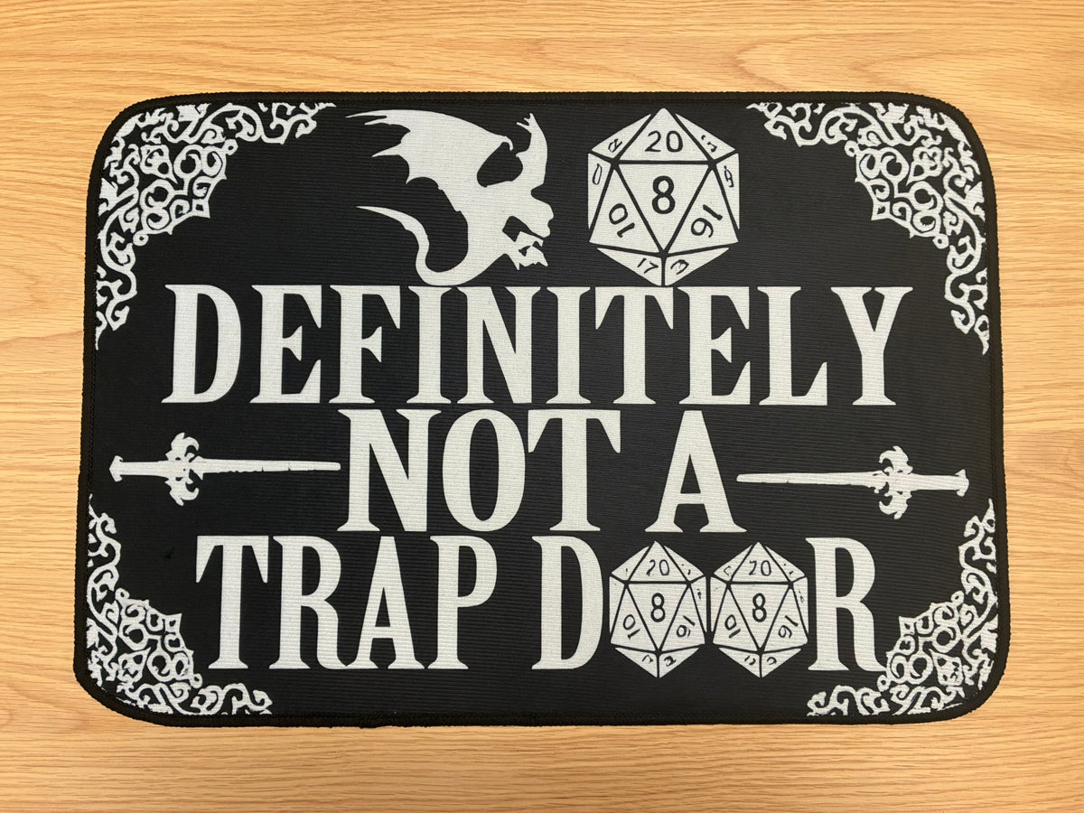 A rectangular black mat with white fantasy-themed text reading 'DEFINITELY NOT A TRAP DOOR' and white decorative corner flourishes, shown on a light wood surface.