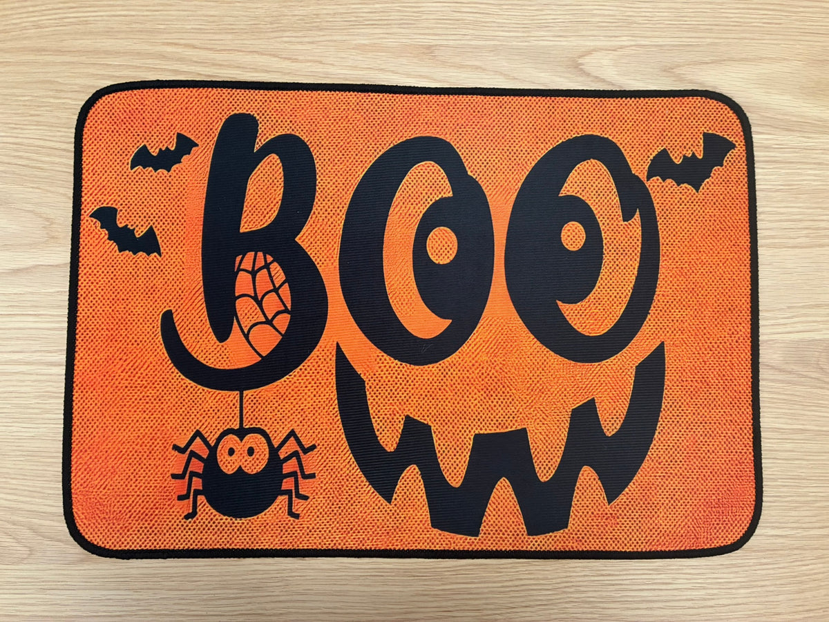 An orange rectangular mat with rounded corners featuring a black Halloween design that spells 'BOO' with a jack-o'-lantern face, spider, bats, and web details, shown on a light wood floor.