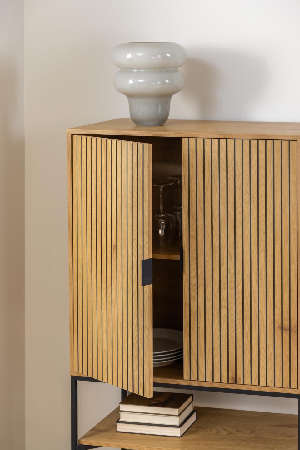 A light oak wood sideboard cabinet with vertical slat doors, shown open with glassware and books inside, topped with a gray ceramic vase against a beige wall.