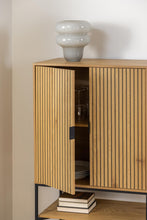 Load image into Gallery viewer, A light oak wood sideboard cabinet with vertical slat doors, shown open with glassware and books inside, topped with a gray ceramic vase against a beige wall.
