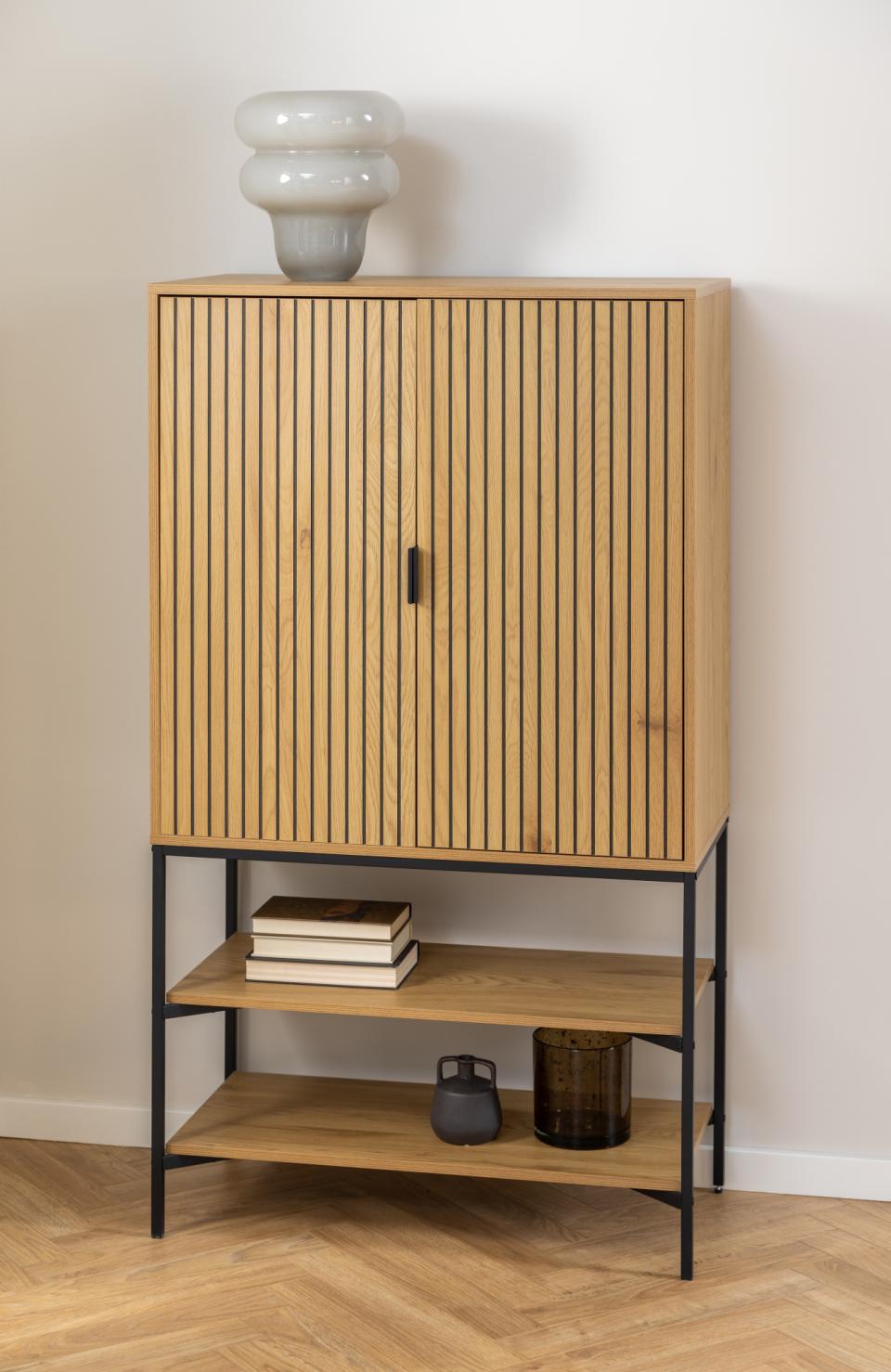 A modern wooden cabinet with a slatted double-door upper section, open lower shelves, and a black metal frame, shown against a plain wall on a wood floor with decorative objects on and around it.