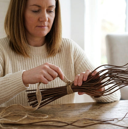 A woman in a cream knit sweater sits at a wooden table, tying a bundle of thin brown twigs together with a piece of natural jute ribbon.