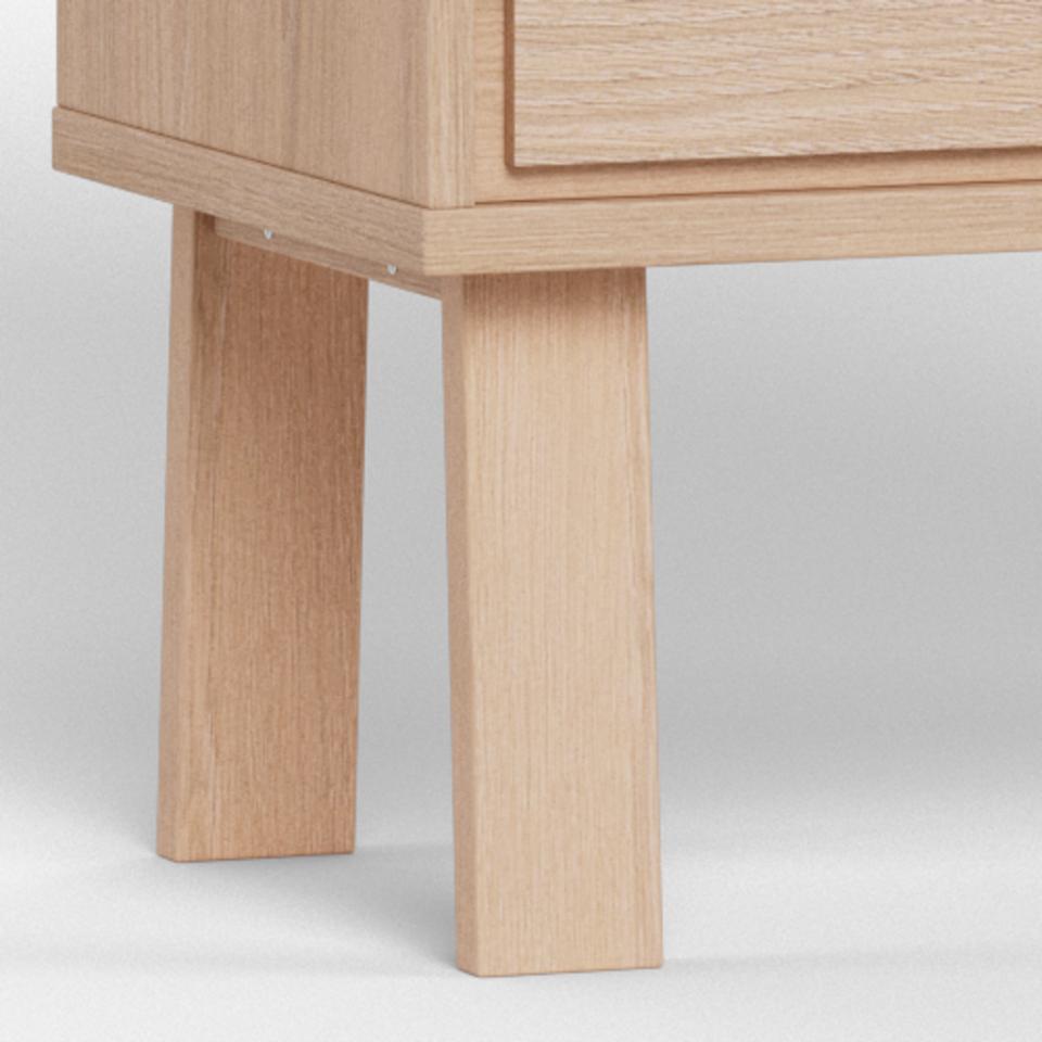Close-up of a light natural oak wood furniture piece showing the front drawer front and two block-style legs on a neutral studio background.
