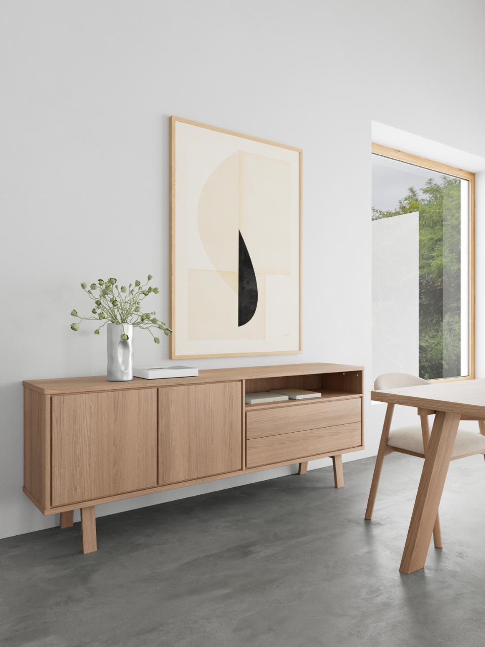 A light oak wood sideboard cabinet styled in a minimalist dining room setting, featuring two cabinet doors, a central drawer, and open shelving. Above it hangs a large abstract art print, and a small vase with green stems sits on top.