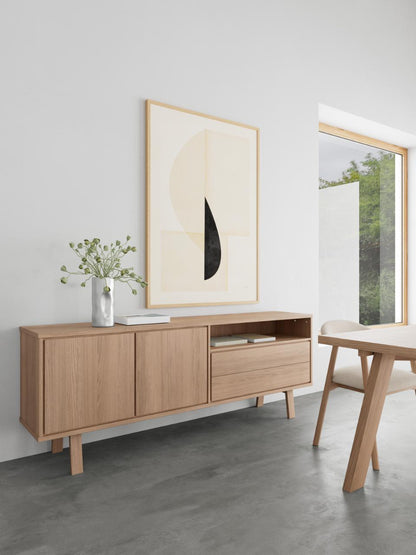 A light oak wood sideboard cabinet styled in a minimalist dining room setting, featuring two cabinet doors, a central drawer, and open shelving. Above it hangs a large abstract art print, and a small vase with green stems sits on top.