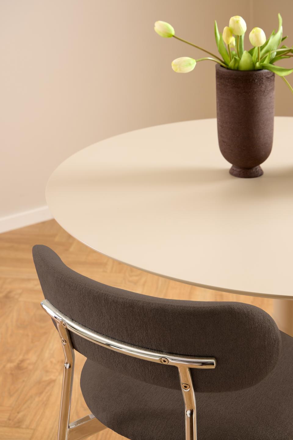 Brown chair in front of a round white table with a vase of yellow tulips on it.