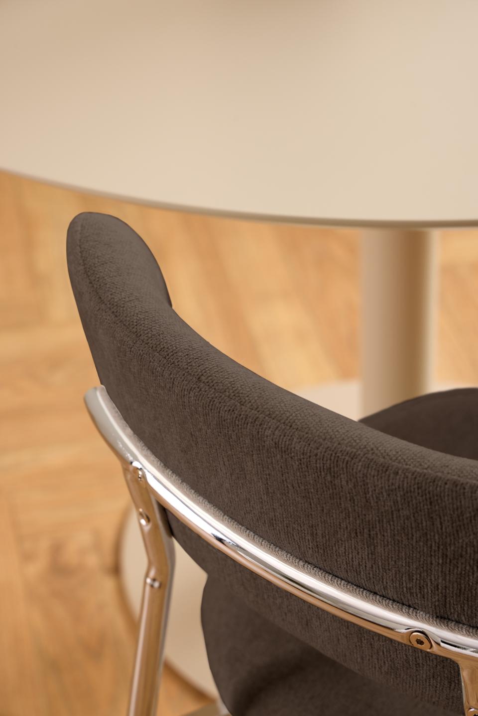 Close-up of a modern chair with dark gray textured fabric upholstery and a curved polished chrome metal frame, set in a warm indoor environment with a light wood floor and a cream-colored table in the background.