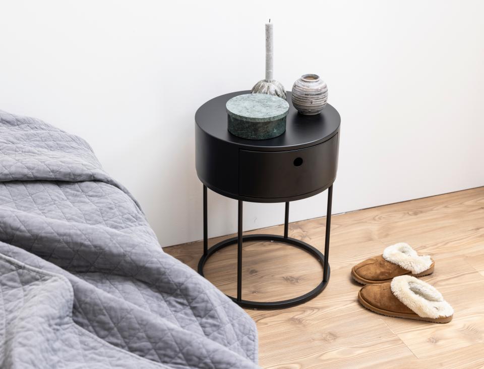 Black round side table with decorative items next to a bed with gray bedding on a wooden floor.