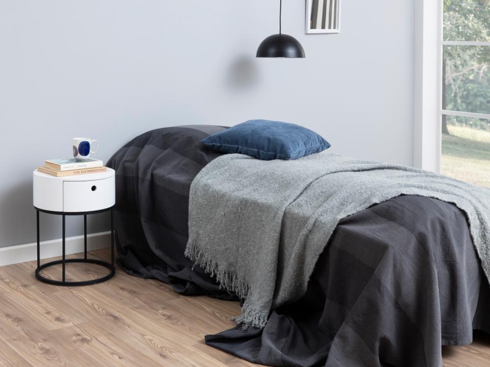 Bedroom with a bed covered in gray and black blankets, a nightstand with a small device, and a lamp.