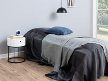 Load image into Gallery viewer, Bedroom with a bed covered in gray and black blankets, a nightstand with a small device, and a lamp.
