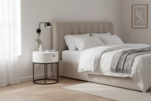 Load image into Gallery viewer, A modern bedroom scene featuring a bed with white linens and a beige upholstered headboard, styled with a white nightstand, black wall lamp, light wood flooring, and sheer curtains.
