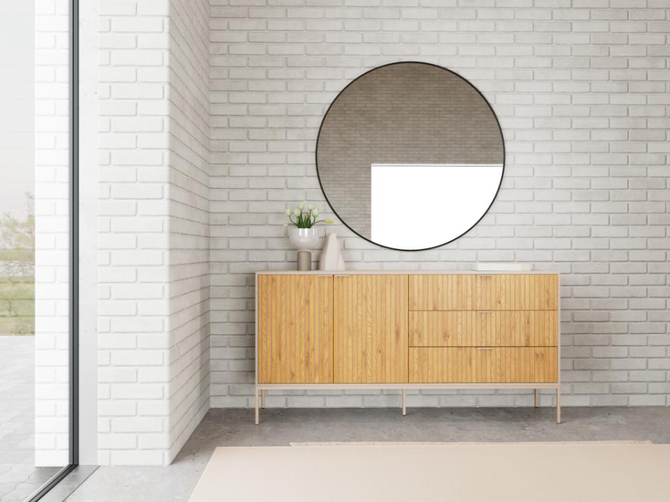 A modern light wood sideboard cabinet with a large round black-framed mirror mounted on a white brick wall above it, set in a minimalist interior with a concrete floor.
