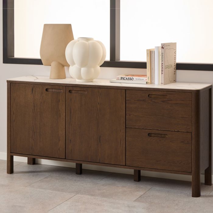 A Southampton modern wooden sideboard with a white marble-look cerammic top, shown in a styled room setting with decorative vases, books, and a window in the background.