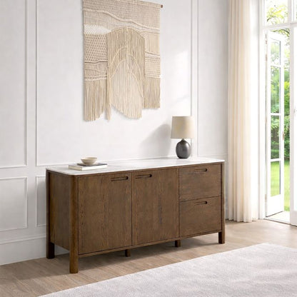 A modern sideboard cabinet with a warm brown wood finish, featuring a white ceramic top, two cabinet doors, and two drawers on the right. It is styled in a bright room with a white marble wall, a woven macrame wall hanging, a black table lamp, and a small bowl.