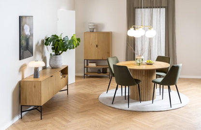 A modern dining room setup featuring a round wooden table with a ribbed pedestal base, surrounded by six dark green upholstered chairs on a round white rug. The room has light wood herringbone flooring, a slatted wood cabinet, and a multi-light pendant chandelier.