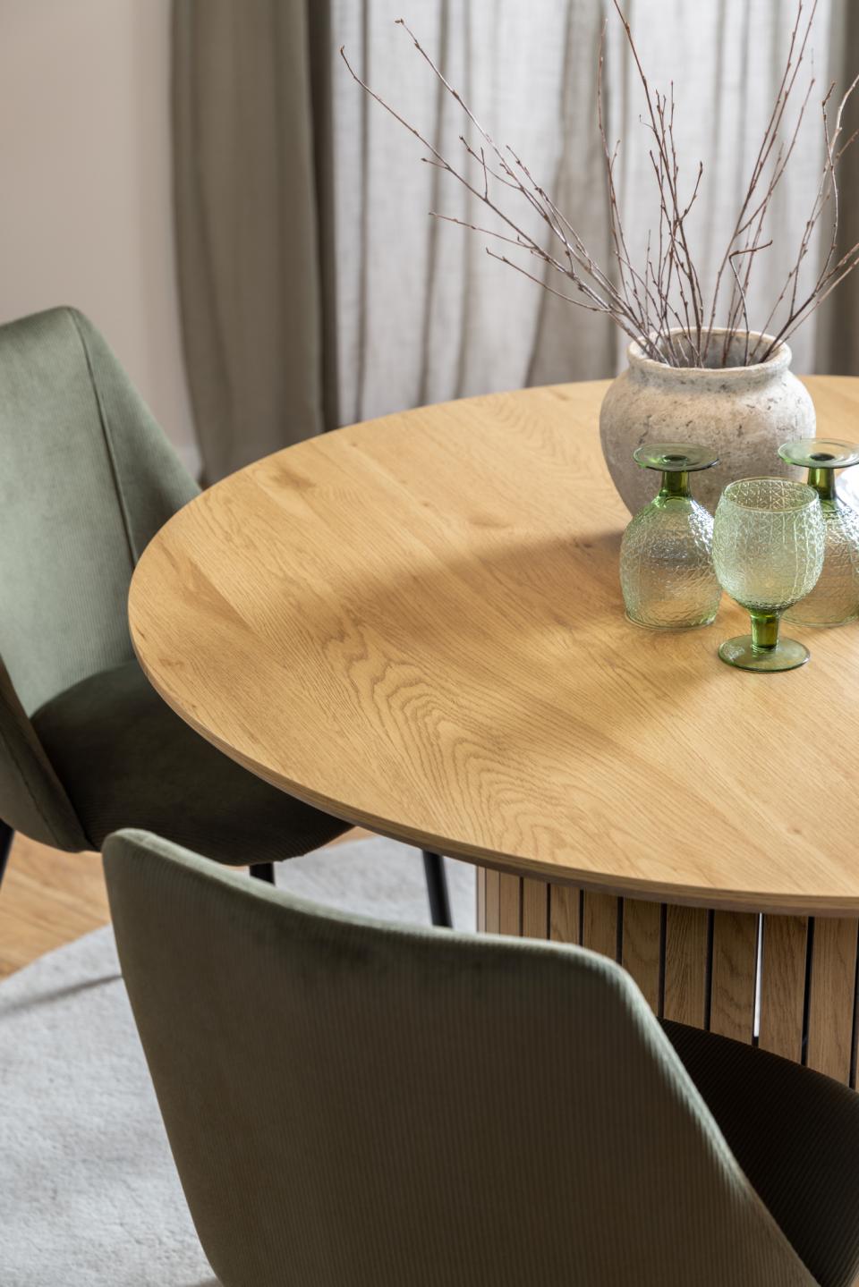 A round light oak wood dining table in a modern dining room setting, styled with a textured ceramic vase containing dried branches and two green glass goblets, next to a green upholstered chair.
